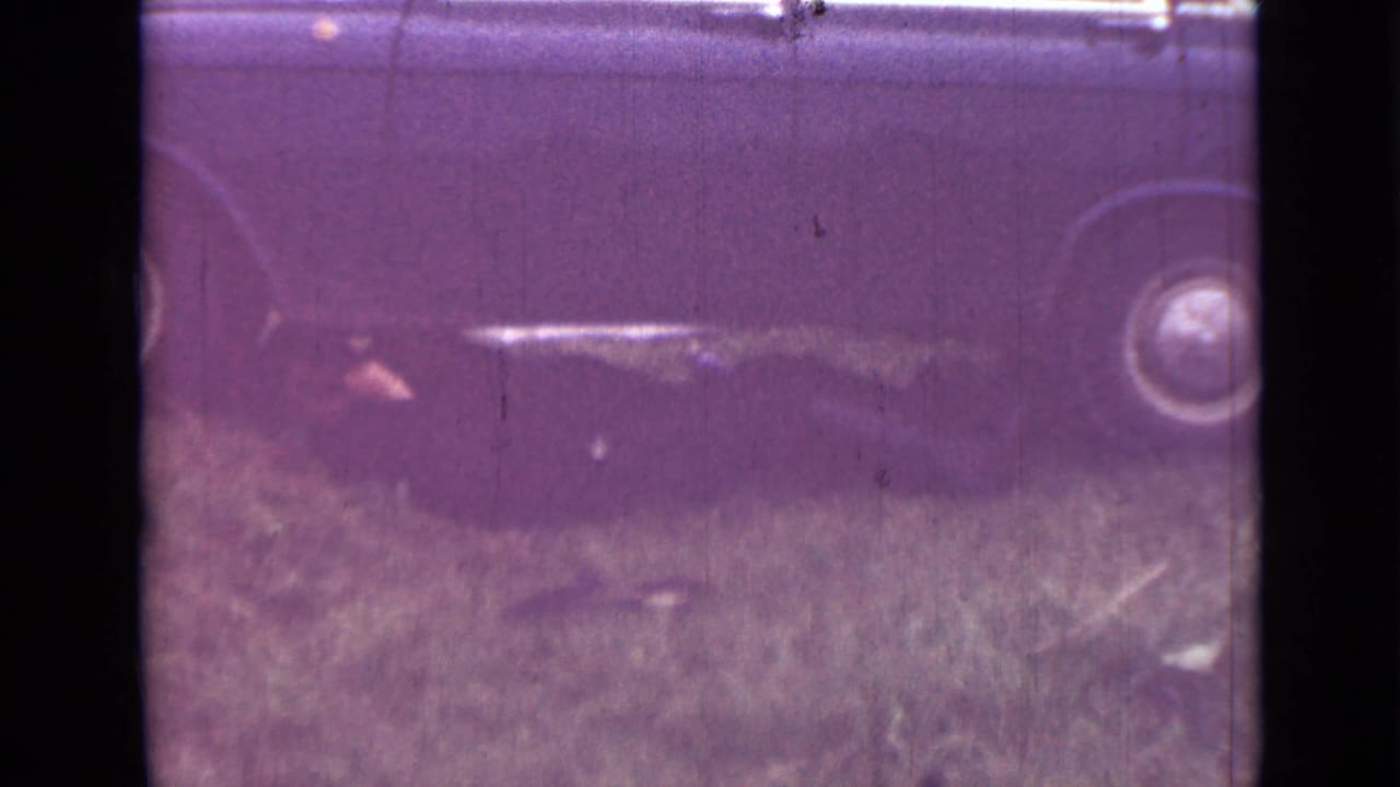 Dark car body over grass with one wheel visible and aged film scratches.