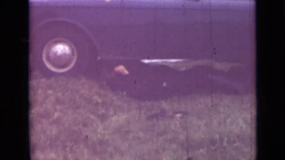 1967 Saint Petersburg home-movie frame showing a close view of a car wheel and side panel above grass, with heavy grain and aged color shift.