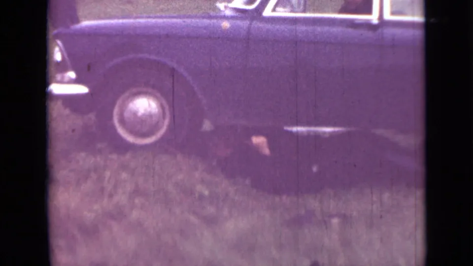 1967 Saint Petersburg 8mm frame showing a dark sedan parked on grass with a person lying partly under the side.