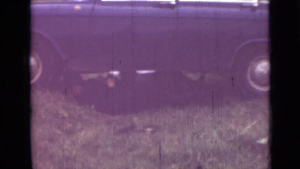 Soft 8mm frame of a parked sedan on uneven grass, with a person lying beneath the vehicle during repairs.