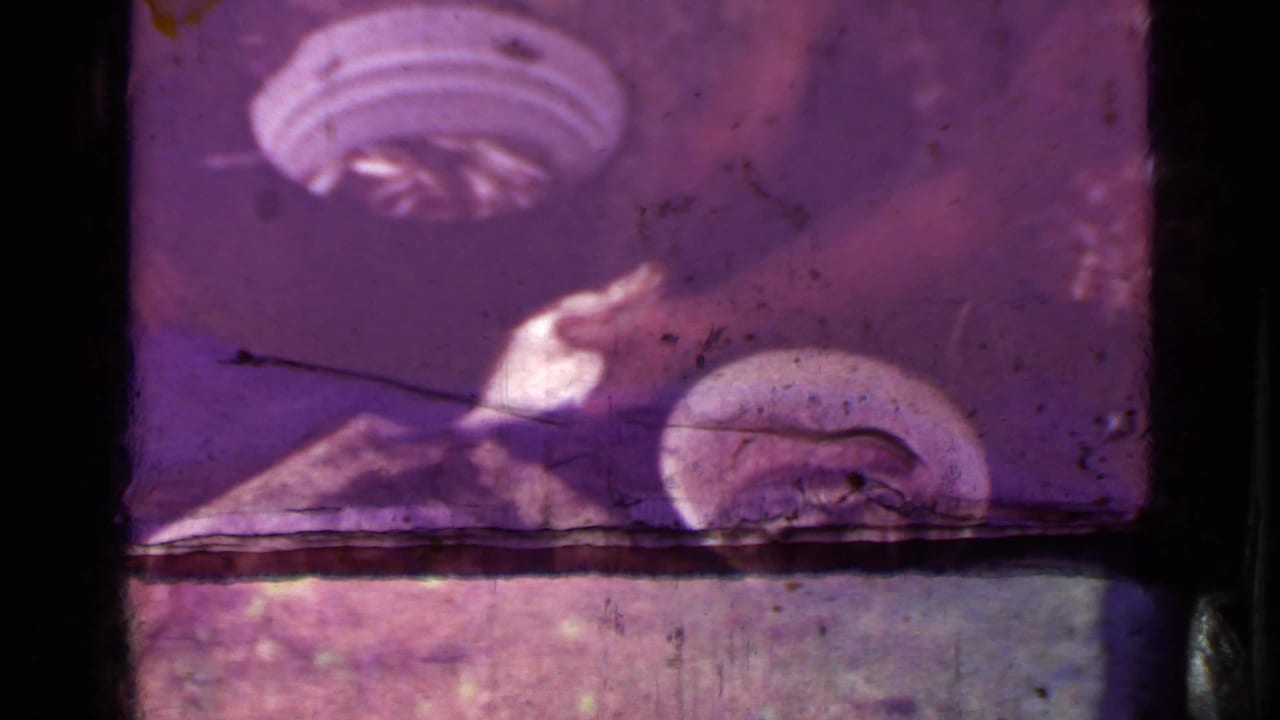 Faded 8mm frame with magenta cast, heavy wear, and indistinct interior-like shapes near a film edge.