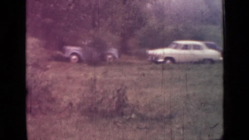 1967 Saint Petersburg home-movie frame showing two parked cars on a grassy lakeside slope with trees behind them.
