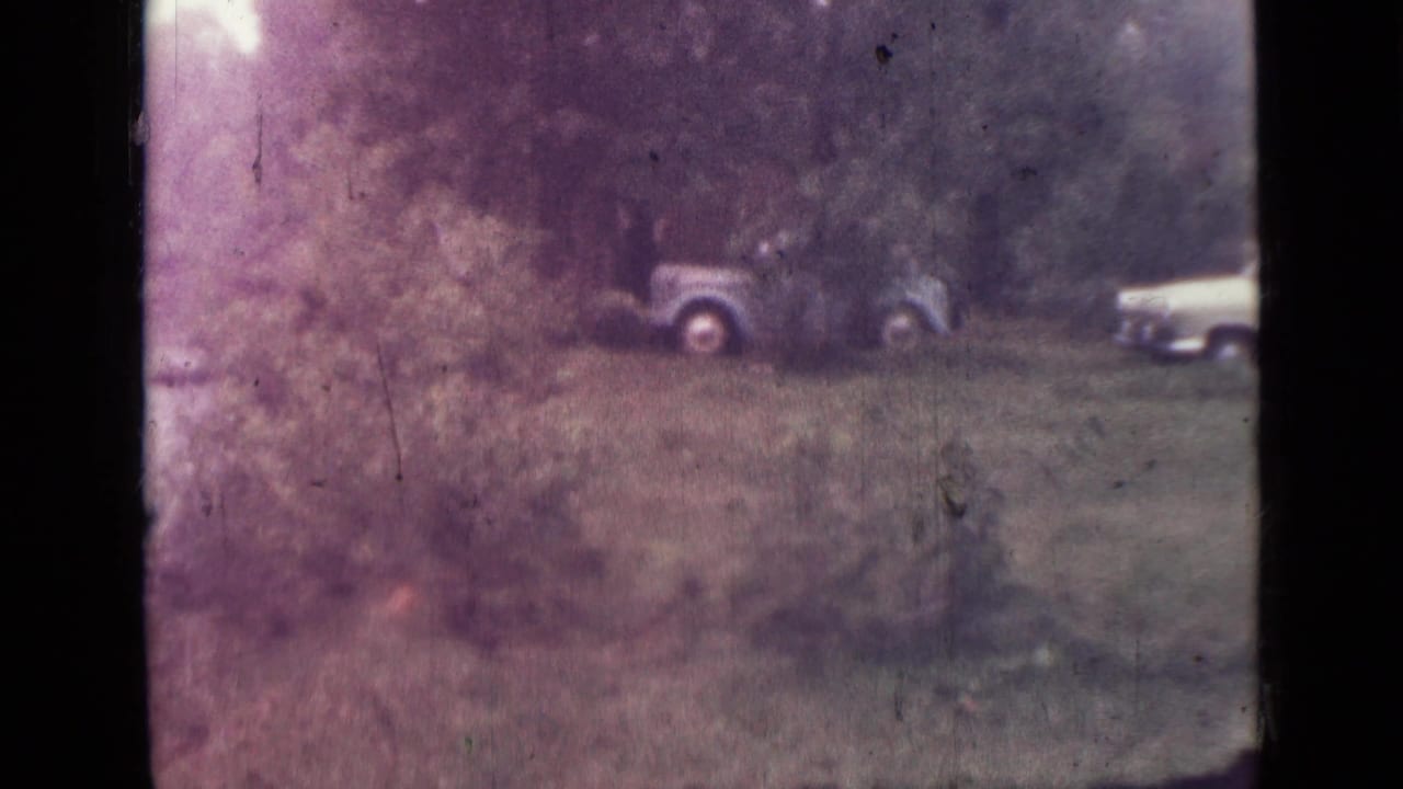 Faded 8mm view of parked mid-century cars near a wooded lake area, with one car partly cropped at the right edge.