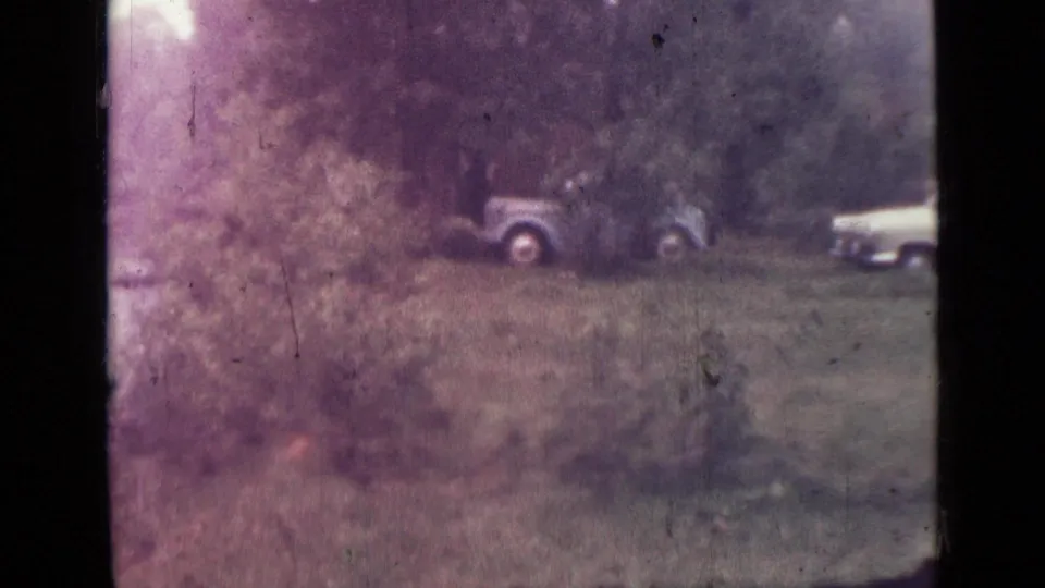 Faded 8mm view of parked mid-century cars near a wooded lake area, with one car partly cropped at the right edge.