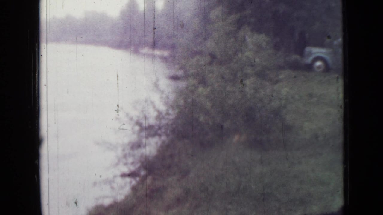 1967 Saint Petersburg lakeside frame with pale water, wooded shoreline, and a parked car partly visible at the right edge.