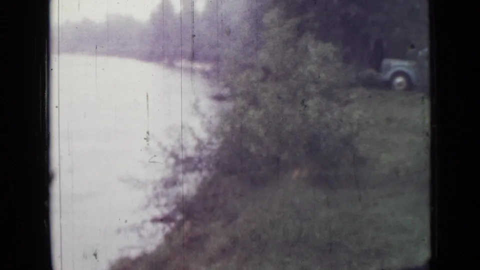 1967 Saint Petersburg lakeside frame with pale water, wooded shoreline, and a parked car partly visible at the right edge.