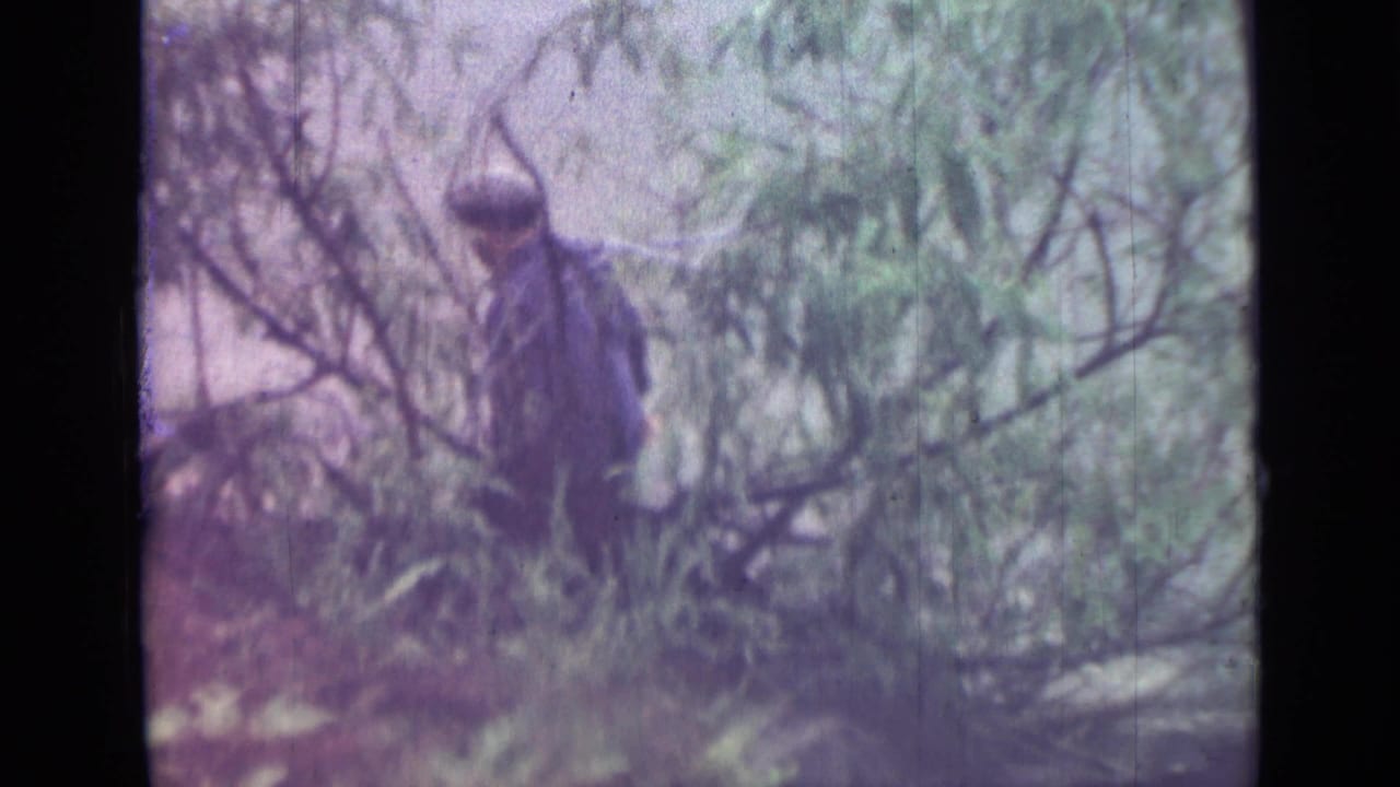 A man in a dark jacket and cap appears partly obscured by brush along the Saint Petersburg riverbank in the 1967 home movie.