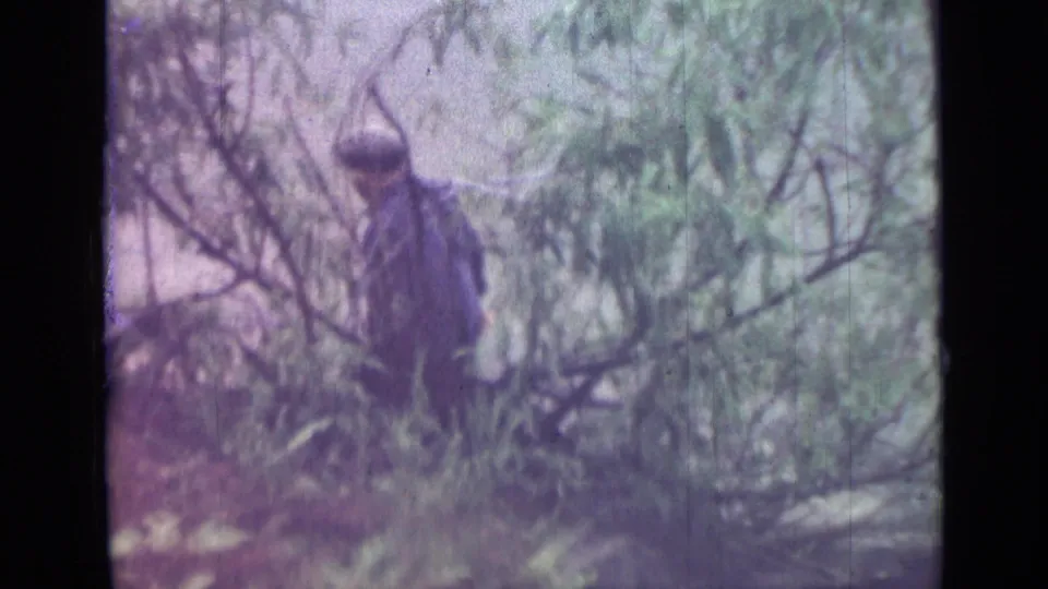 A man in a dark jacket and cap appears partly obscured by brush along the Saint Petersburg riverbank in the 1967 home movie.