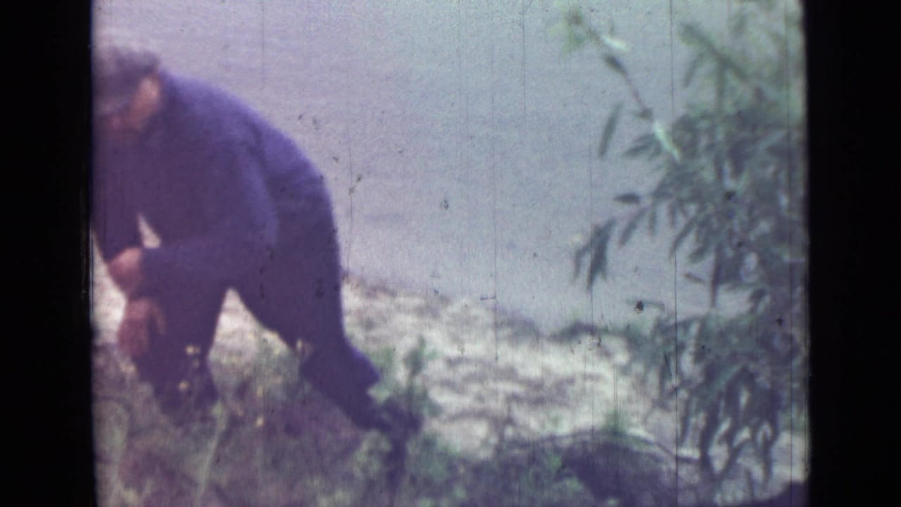 In 1967 Saint Petersburg home-movie footage, a man in a cap and dark work clothes bends near low greenery on a small slope.