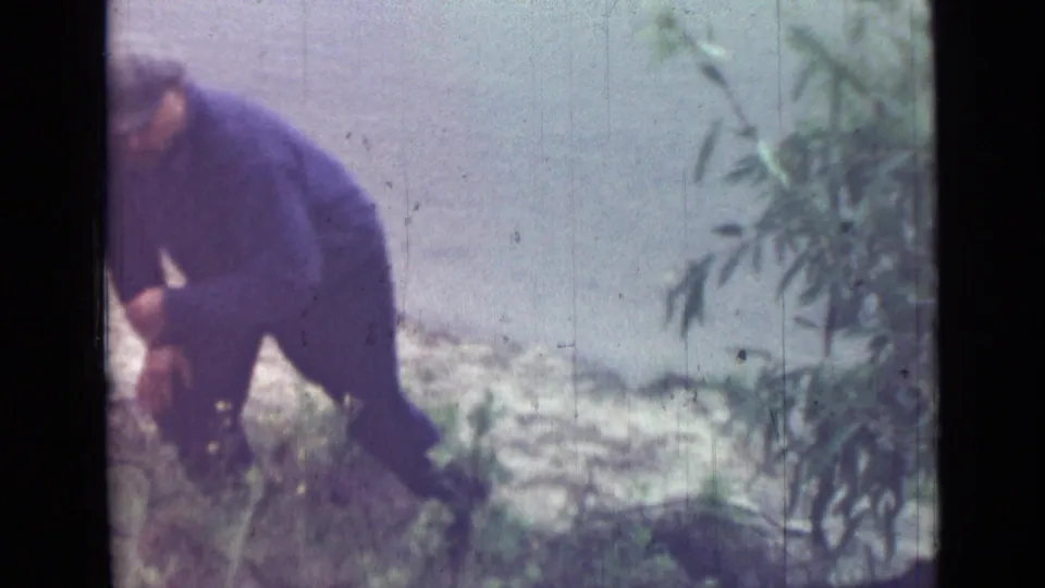 In 1967 Saint Petersburg home-movie footage, a man in a cap and dark work clothes bends near low greenery on a small slope.