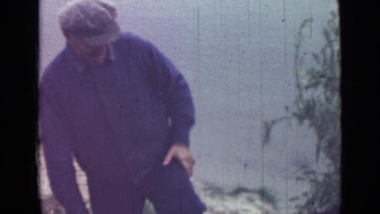 The man leans forward in dark workwear, with low plants at the edge of the frame and a pale waterside or open background behind him.