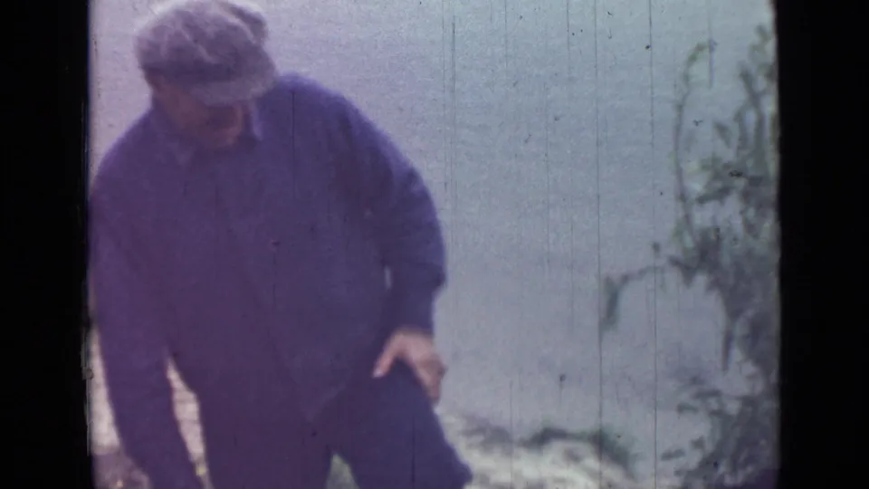 The man leans forward in dark workwear, with low plants at the edge of the frame and a pale waterside or open background behind him.