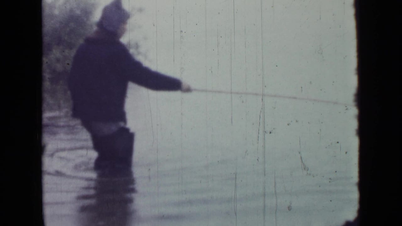 1967 8mm home-movie frame showing a person in dark clothing and boots fishing from shallow lake water.