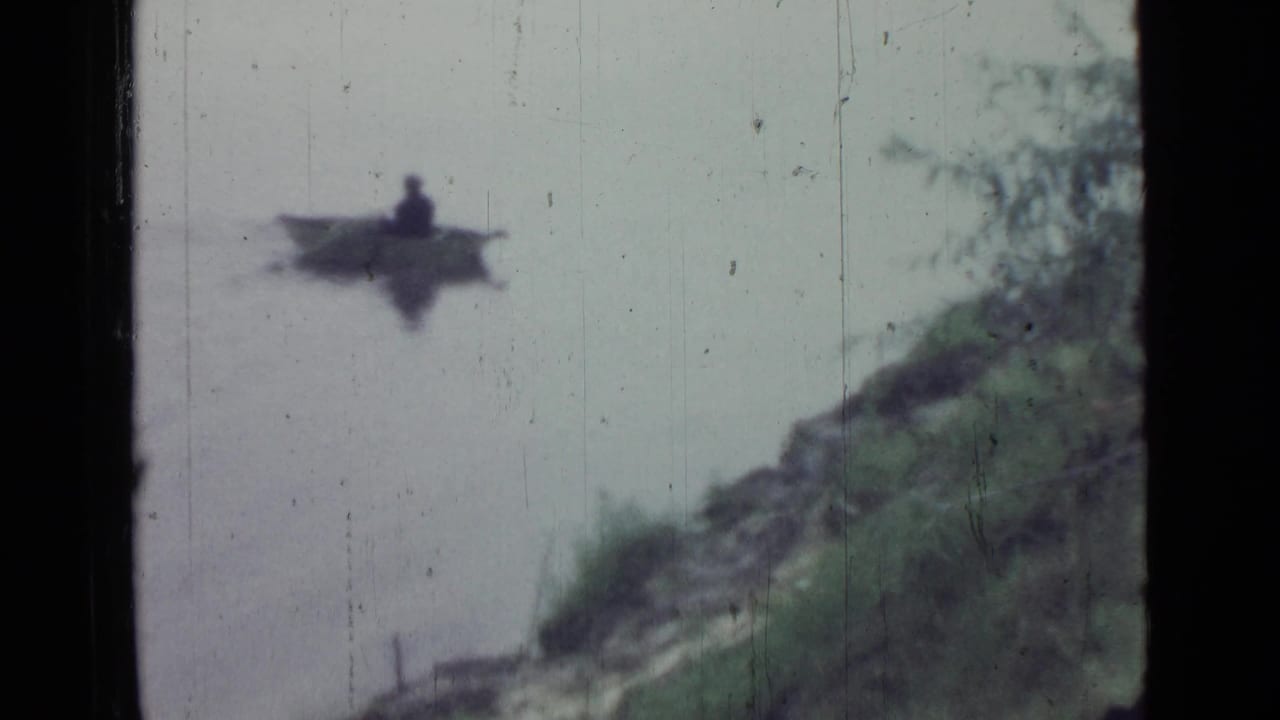 A small rowboat with a seated person moves across calm water toward a grassy bank in the 1967 home-movie scene.