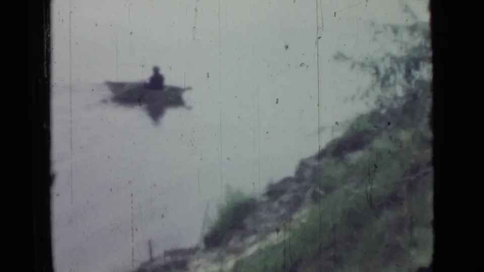 A small rowboat with a seated person moves across calm water toward a grassy bank in the 1967 home-movie scene.