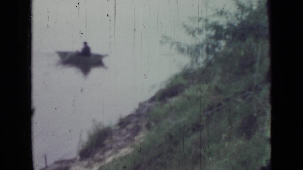 The rowboat continues across pale lake water, seen beyond a sloped grassy shoreline in archival 8mm footage.