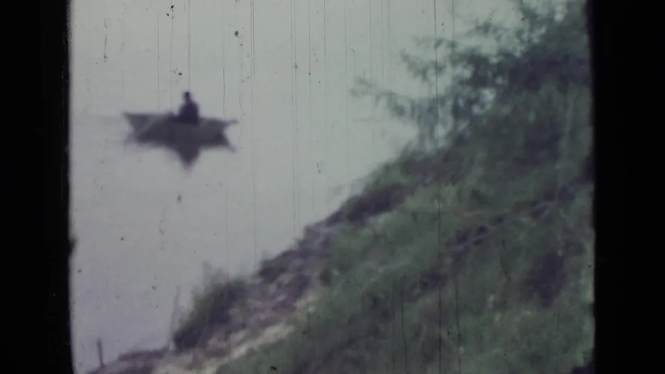 The rowboat continues across pale lake water, seen beyond a sloped grassy shoreline in archival 8mm footage.