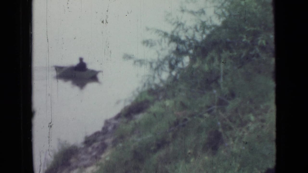 A distant seated person in a small rowboat appears near the grassy shore, with soft focus and visible 8mm film wear.
