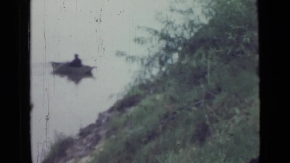 A distant seated person in a small rowboat appears near the grassy shore, with soft focus and visible 8mm film wear.