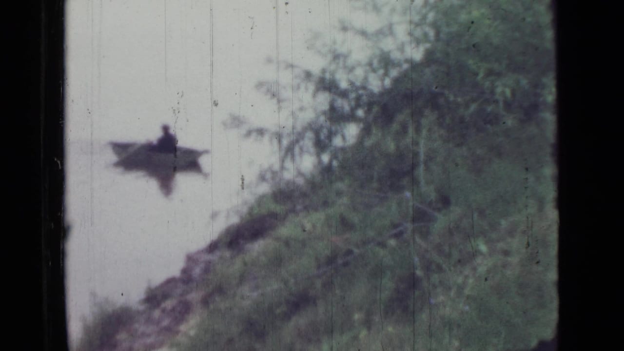 1967 Saint Petersburg 8mm frame showing a lone person rowing a small boat near a tree-lined embankment on still water.