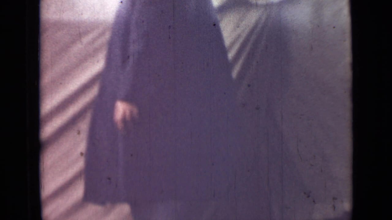 The lower portion of the man in a long dark coat is visible near pale canvas, with one hand and dark shoes in frame.