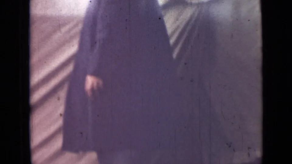 The lower portion of the man in a long dark coat is visible near pale canvas, with one hand and dark shoes in frame.
