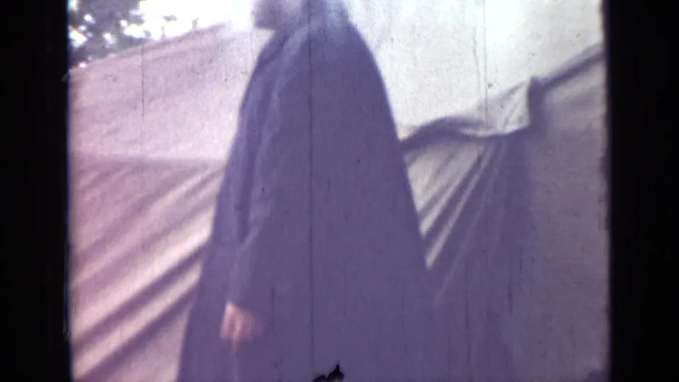 The man in a long dark coat walks past pale canvas tenting in the stated 1967 Saint Petersburg home-movie clip.