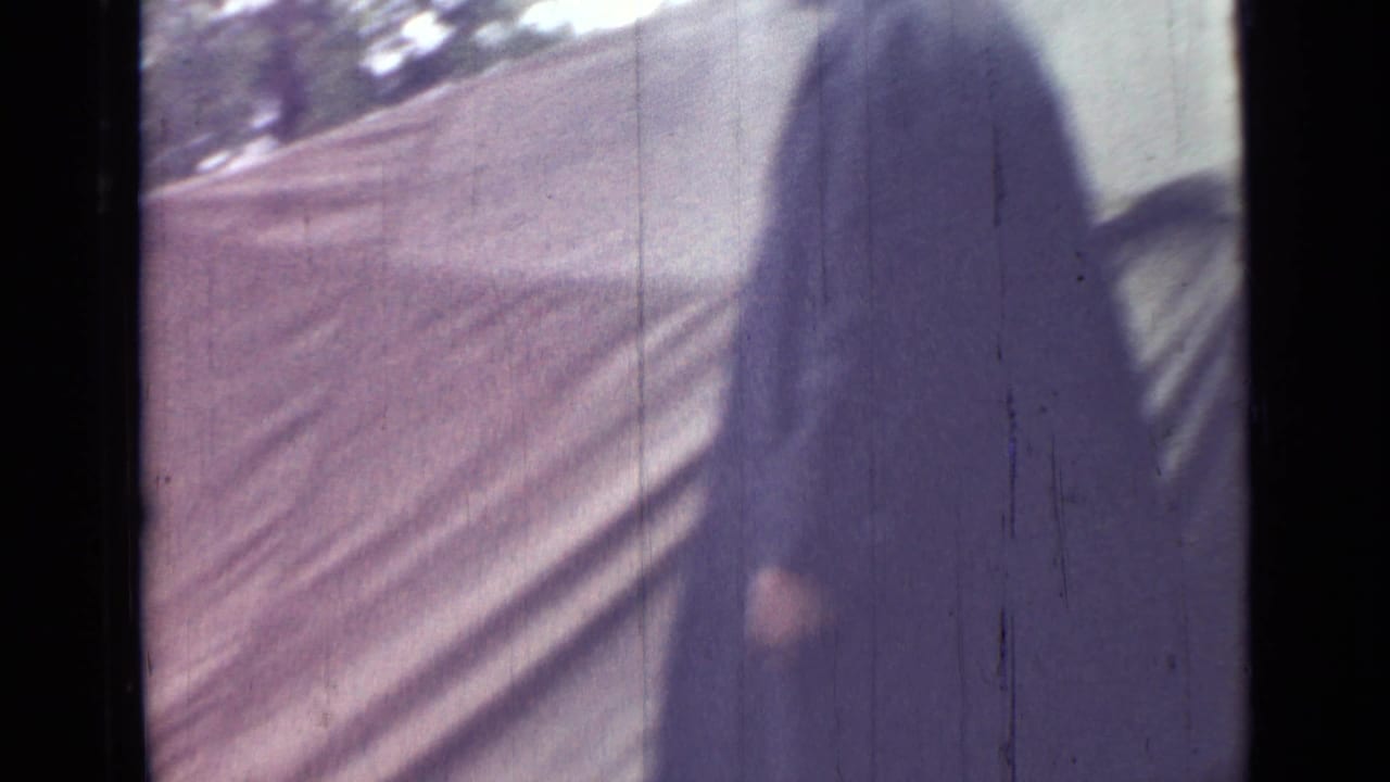 Person in a dark coat walking beside a sloped canvas tent in a wooded campsite.