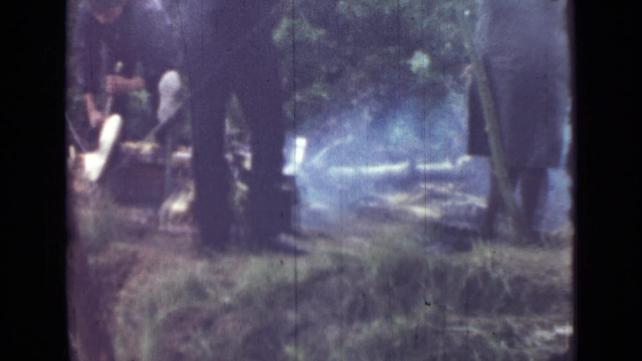 1967 8mm frame near Saint Petersburg showing adults around a small smoking campfire in a wooded campsite.