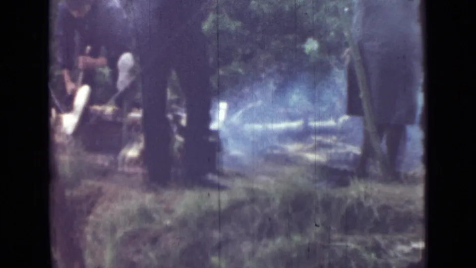 1967 8mm frame near Saint Petersburg showing adults around a small smoking campfire in a wooded campsite.