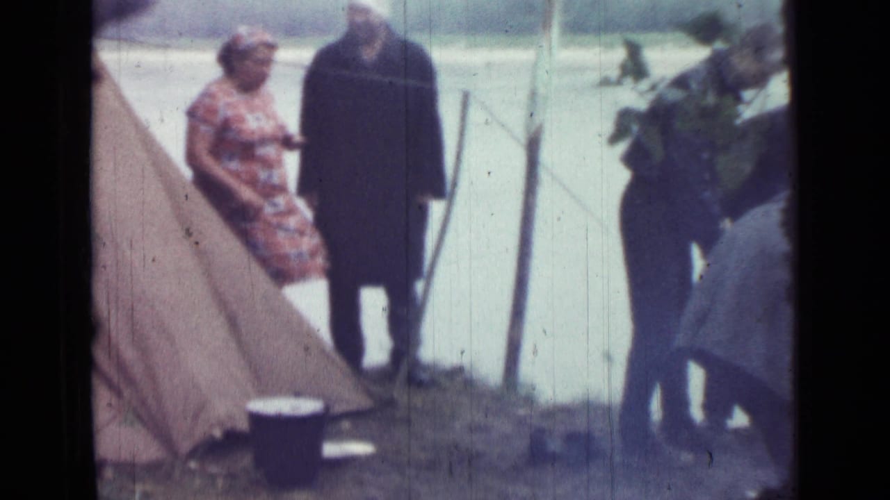 1967 Saint Petersburg home-movie frame showing adults near a canvas tent by the river, including a woman in a patterned dress and a man in a dark coat.