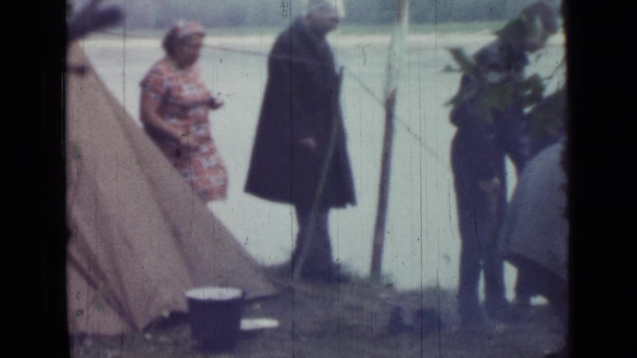 A woman and man stand near a tent while another adult works beside plants at the riverbank.