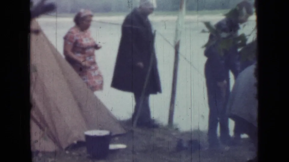 1967 Saint Petersburg riverside camping scene with a woman near the tent, a man in a dark coat, and another adult handling plants or gear.