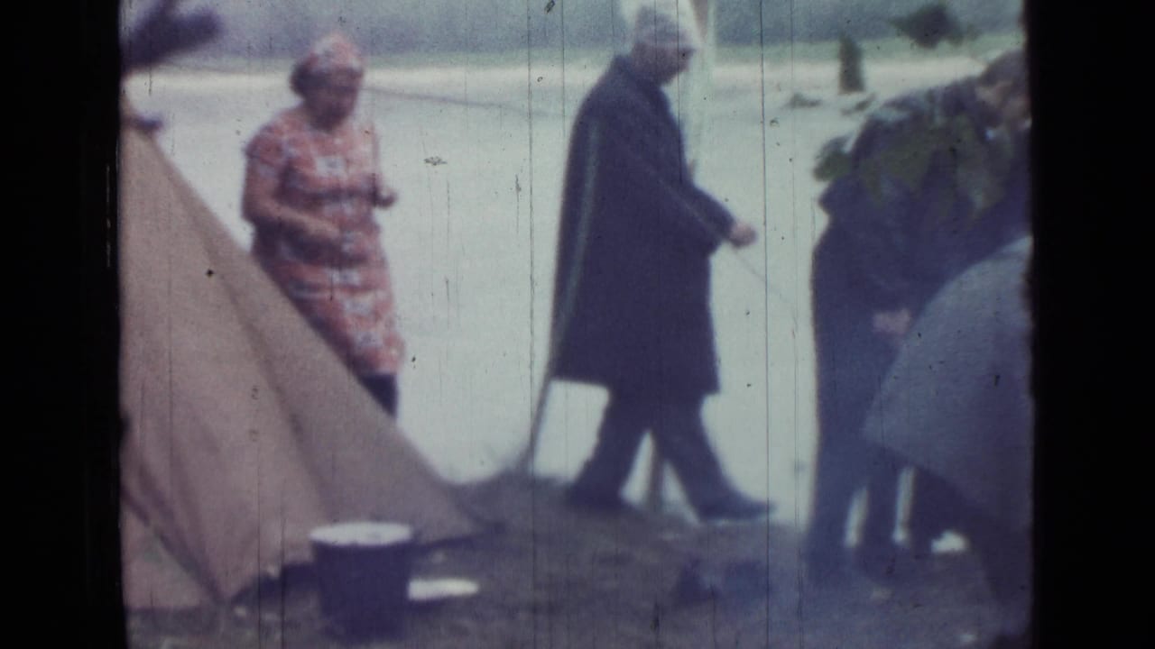 1967 Saint Petersburg home-movie still of adults moving around a riverside campsite with a canvas tent, poles, and bucket visible.
