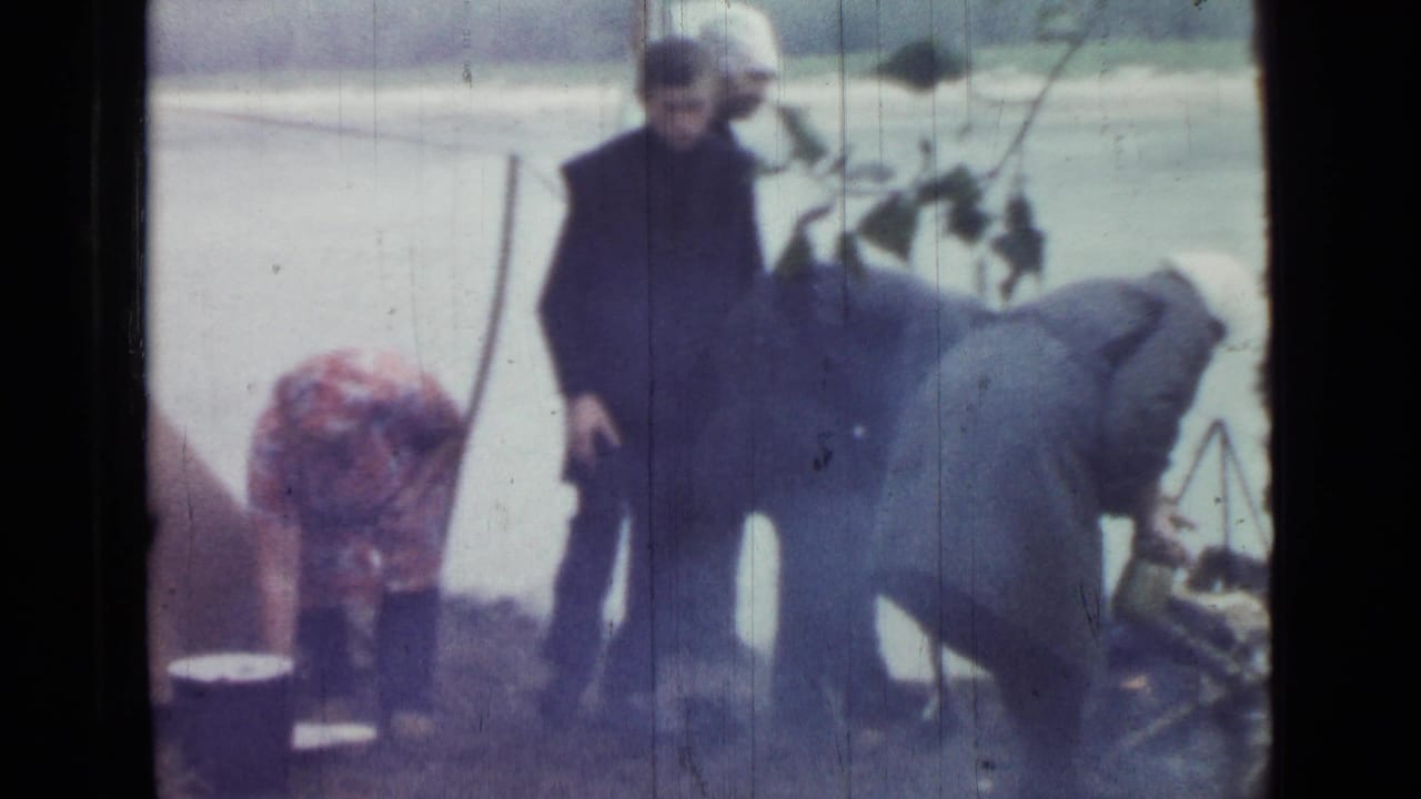 Three adults stand near a lakeside tent and cooking area in soft 8mm footage.
