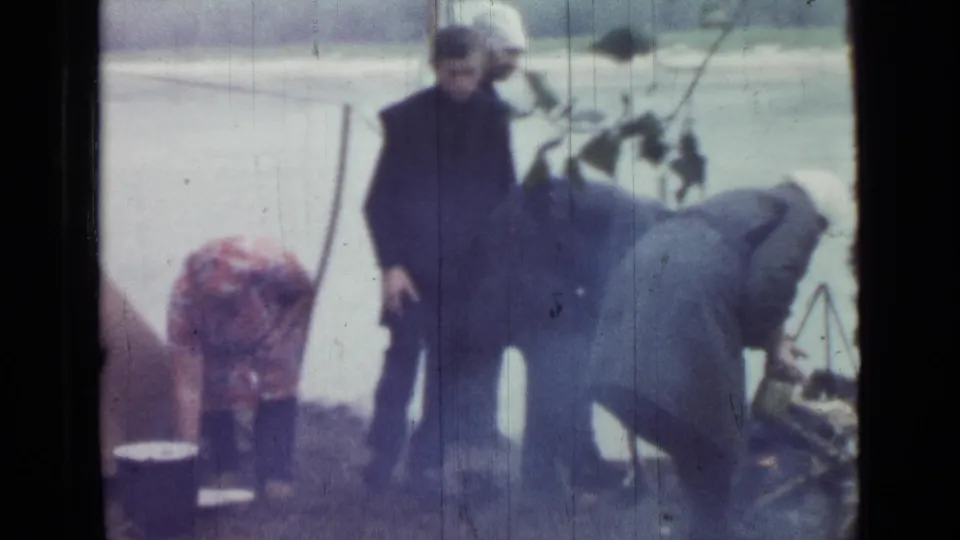 1967 home-movie frame from Saint Petersburg, Russia, showing adults near a tent, water, and a small camp cooking setup.