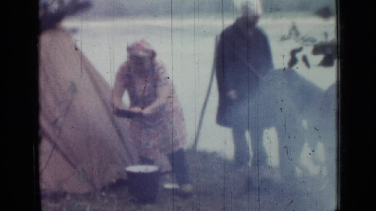 1967 Saint Petersburg home-movie frame showing a woman tending a pot near a tent while another adult stands nearby.