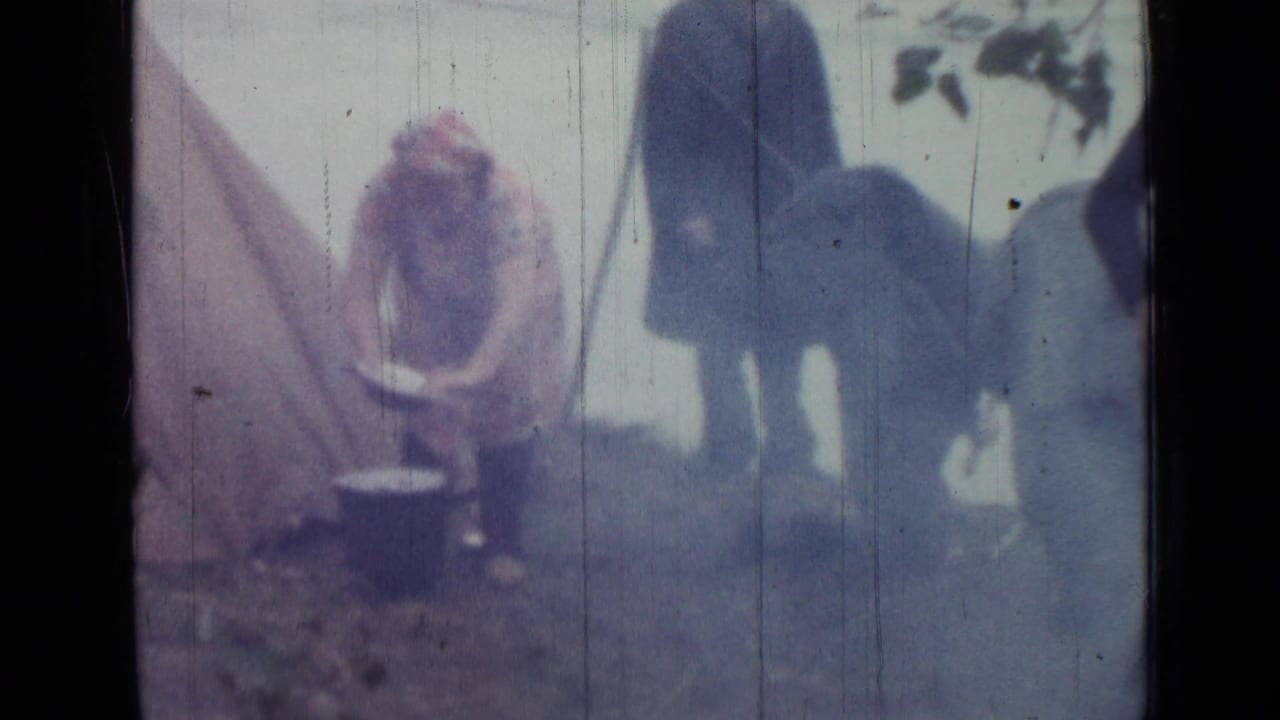 Woman bends over cooking pots beside a tent while two adults stand nearby.
