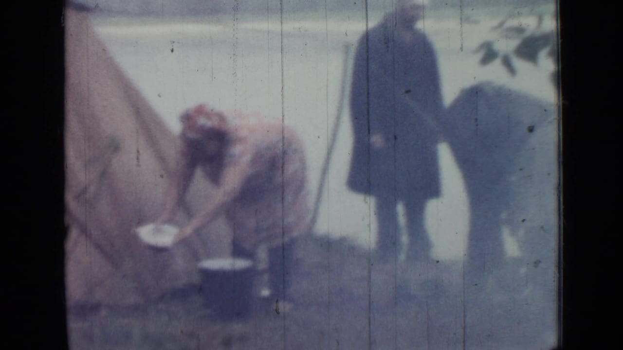 1967 Saint Petersburg camping frame with a woman tending pots beside a tent and adults standing nearby by the water.