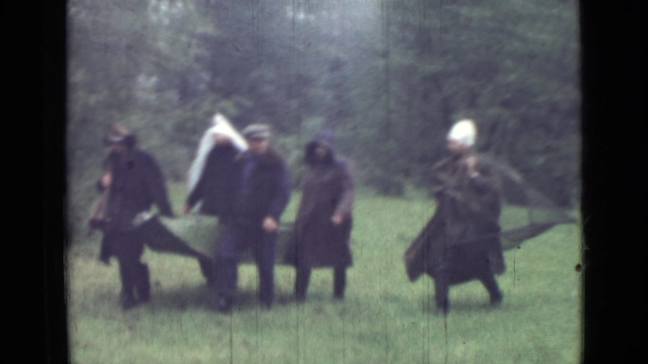 In soft 8mm footage, adults in dark coats move across grass with a stretcher-like carrier and wooded background.