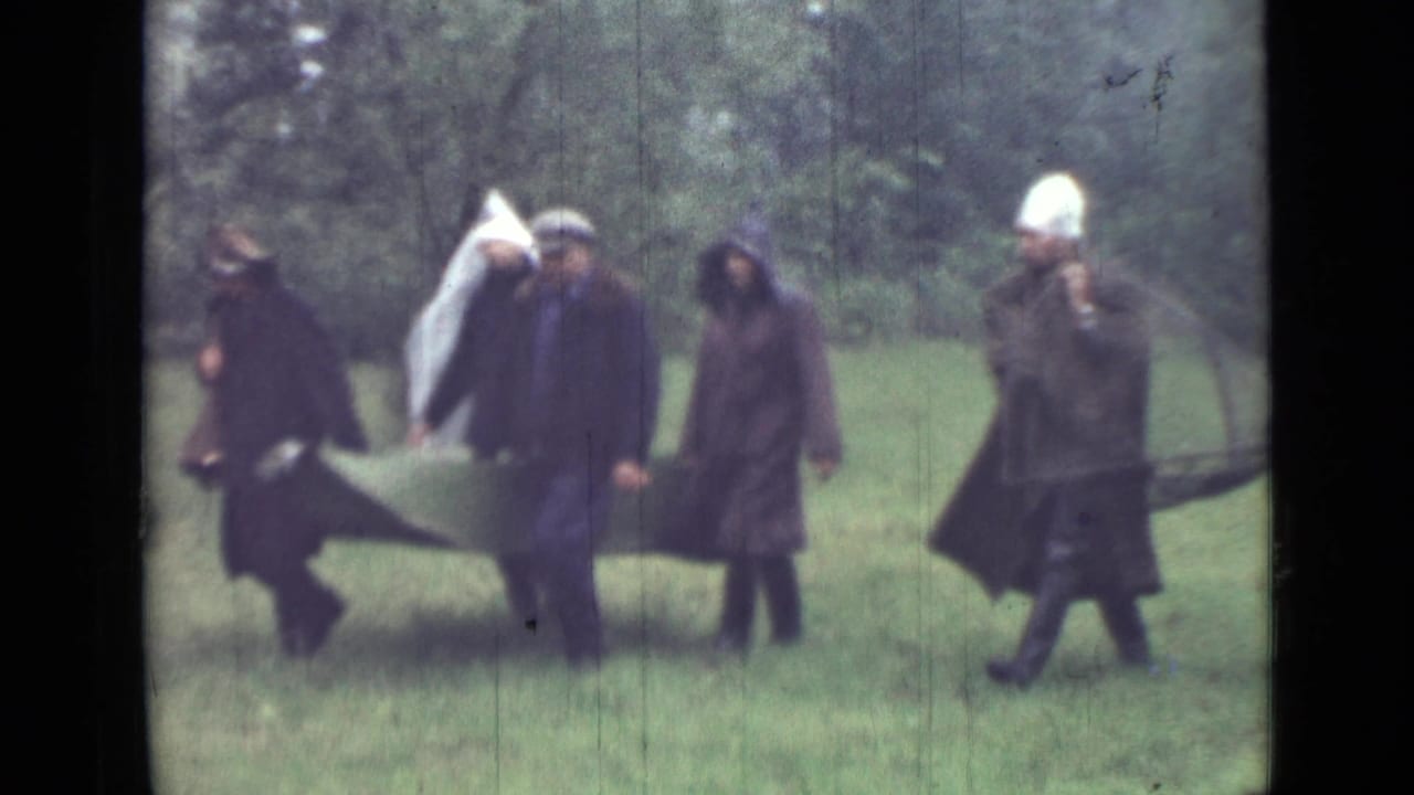1967 Saint Petersburg home-movie frame with adults in dark coats beside a canvas stretcher-like carrier in a grassy wooded area.
