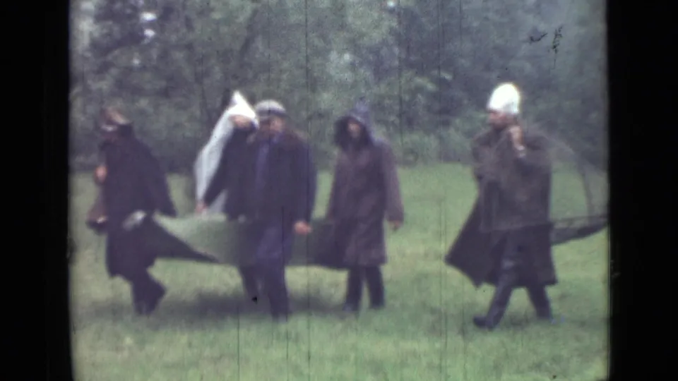 1967 Saint Petersburg home-movie frame with adults in dark coats beside a canvas stretcher-like carrier in a grassy wooded area.