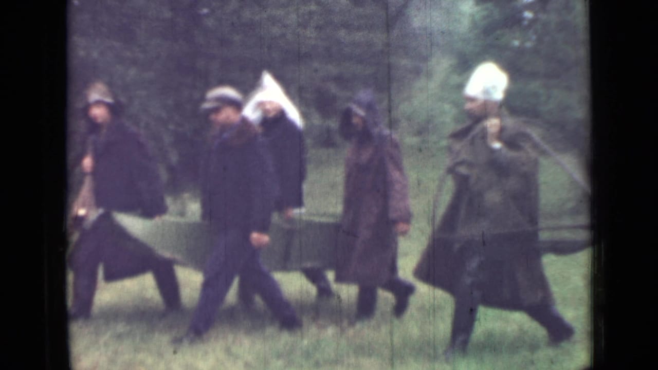 A group of men passes through grass with green canvas and a white cloth visible.