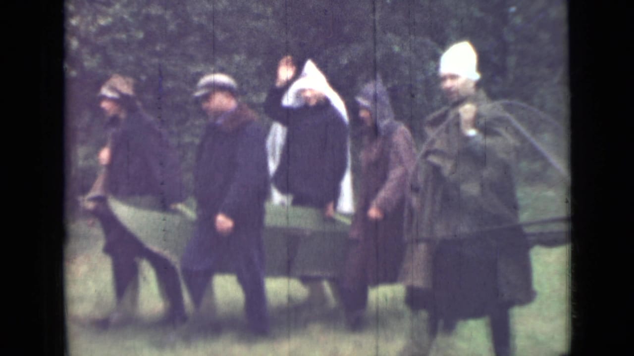 The 1967 home-movie frame shows men in dark coats and hats moving with canvas sheets across a grassy wooded setting.