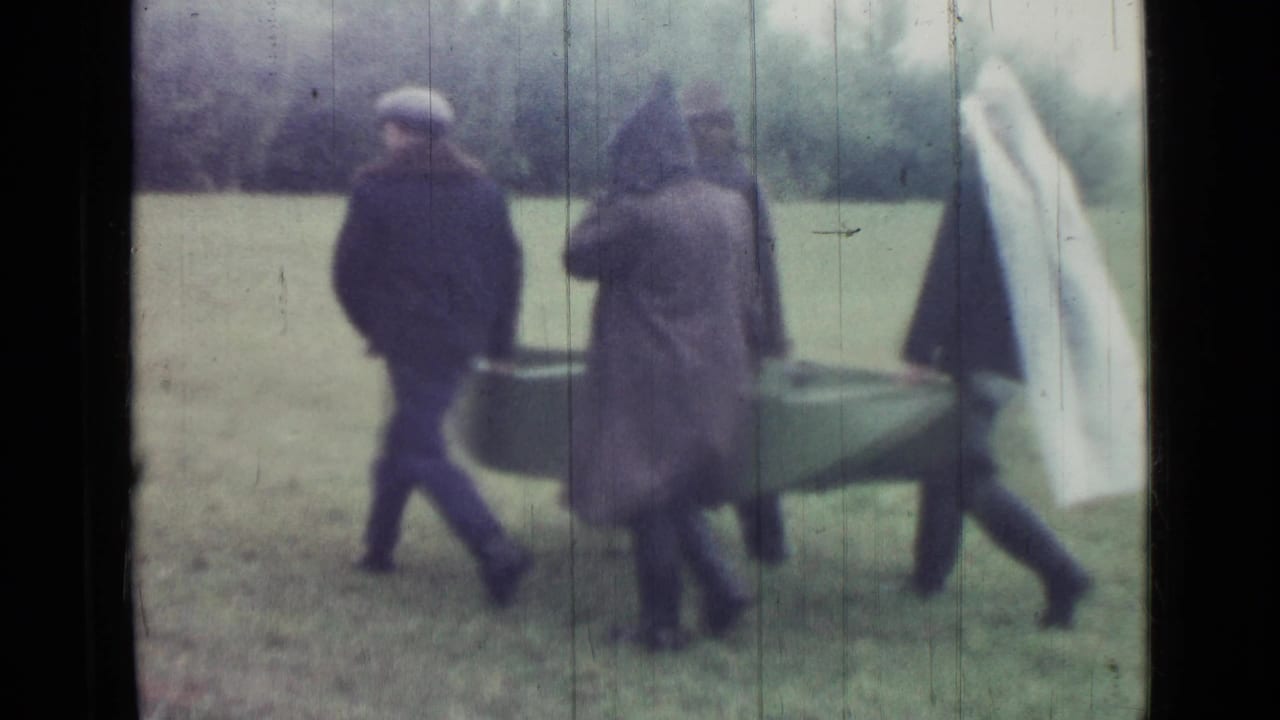 Men in raincoats carry a small boat across a grassy field.