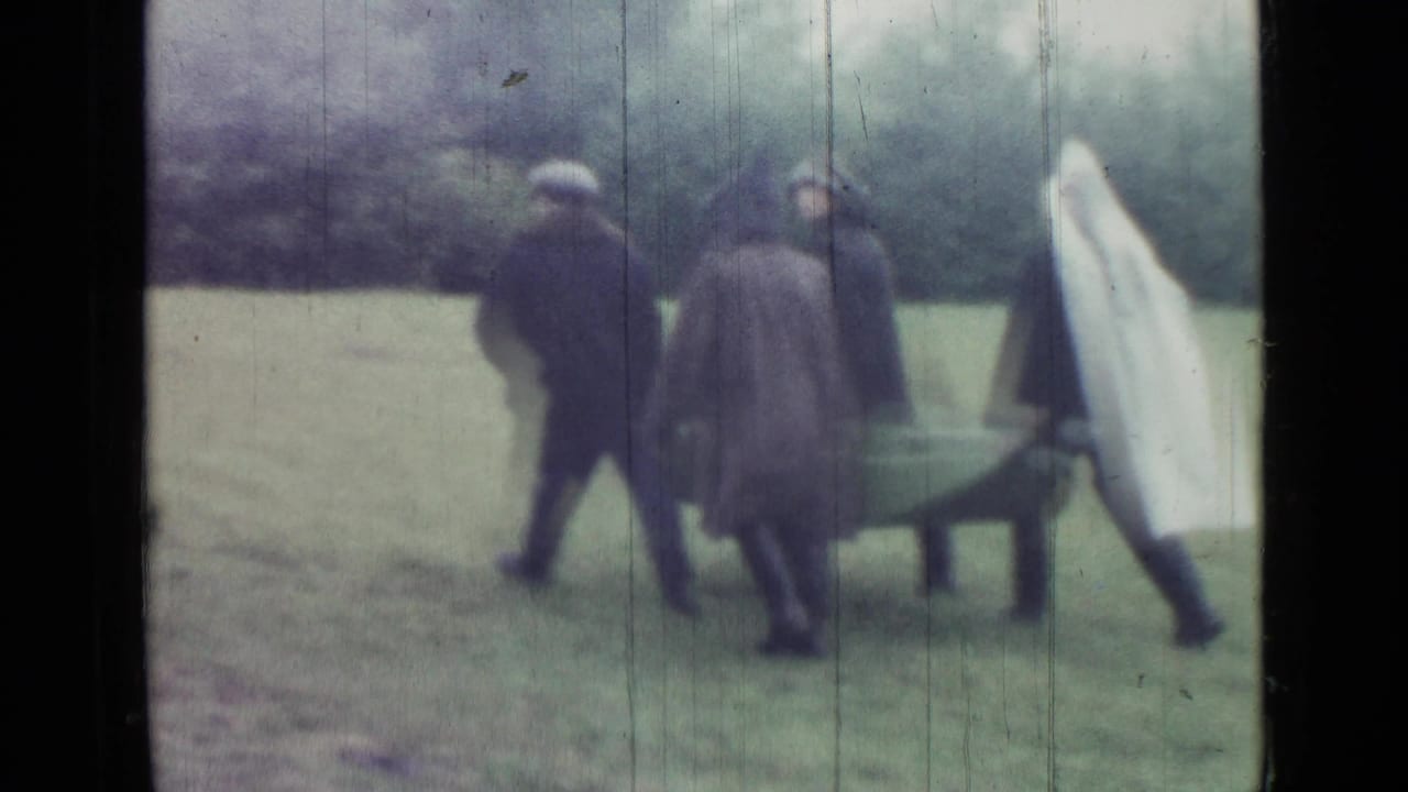 Men in dark raincoats move a small boat across grass toward a wetland area, with a white cover at the side.