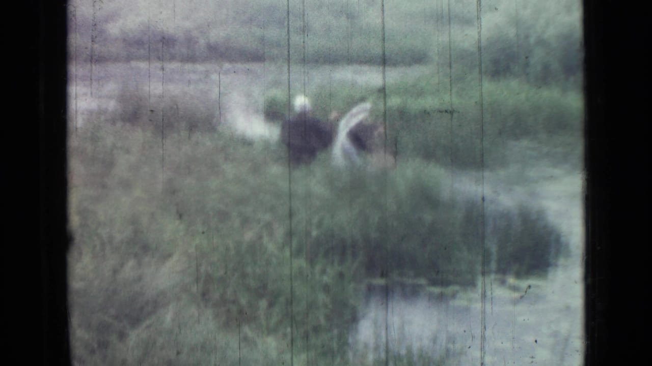 1967 Saint Petersburg 8mm frame showing two distant people in dark outerwear near reeds and water, with soft focus and film scratches.