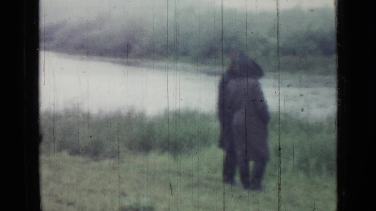 Hooded figure in a dark coat stands near open water and grass in a 1967 Saint Petersburg 8mm home-movie frame.