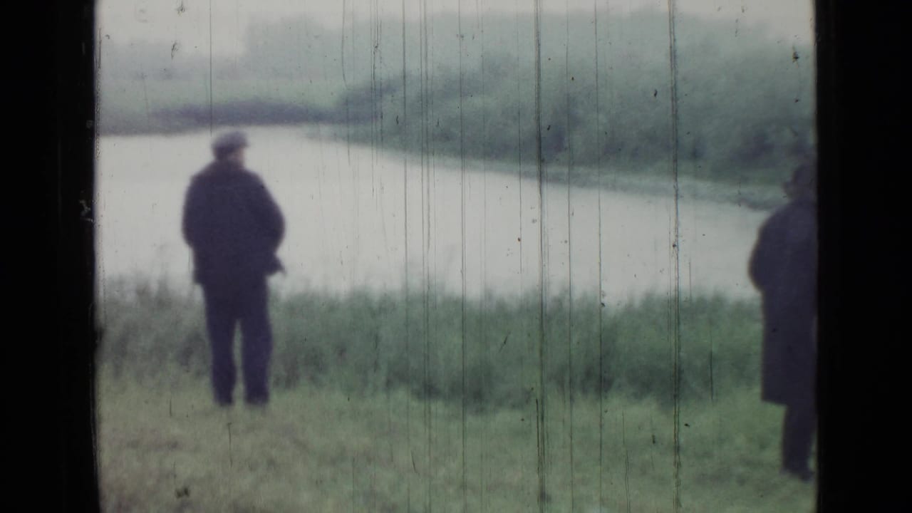 Adult in a dark coat stands near a misty lake with another figure partly visible at the right edge.