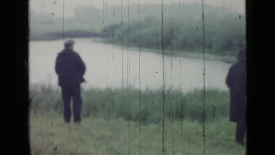 1967 Saint Petersburg home-movie frame showing adults in dark coats near a lake or riverbank, seen through dark vertical framing.